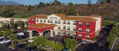 A multi-story hotel with a red and white facade, a parking lot, and a hilly landscape in the background.