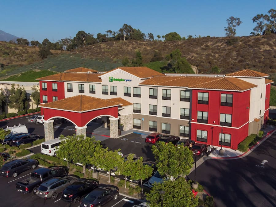 Un hotel de varios pisos con fachada roja y blanca, un estacionamiento y un paisaje montañoso al fondo.