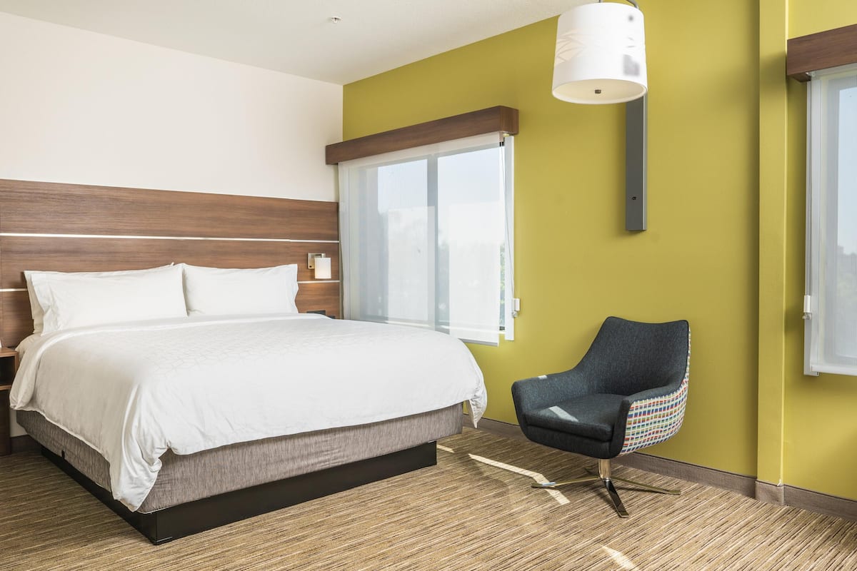 A hotel room with a large bed, a chair, a window, and a wall-mounted lamp.