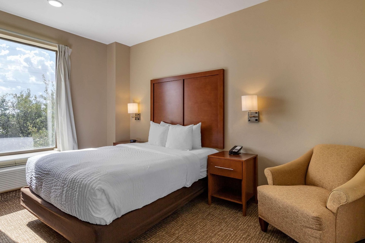 A hotel room with a large bed, a wooden headboard, two bedside lamps, a chair, and a window with curtains.