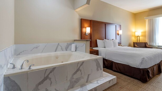 Ein Hotelzimmer mit einer großen Badewanne, einem Bett mit weißer Bettwäsche, einem hölzernen Kopfteil und einem Nachttisch mit einer Lampe.