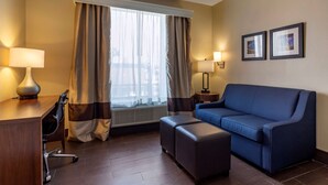 Ein Hotelzimmer mit einem blauen Sofa, einem Schreibtisch mit Lampe, einem Sessel, einem Fenster mit Vorhängen und zwei gerahmten Bildern an der Wand.