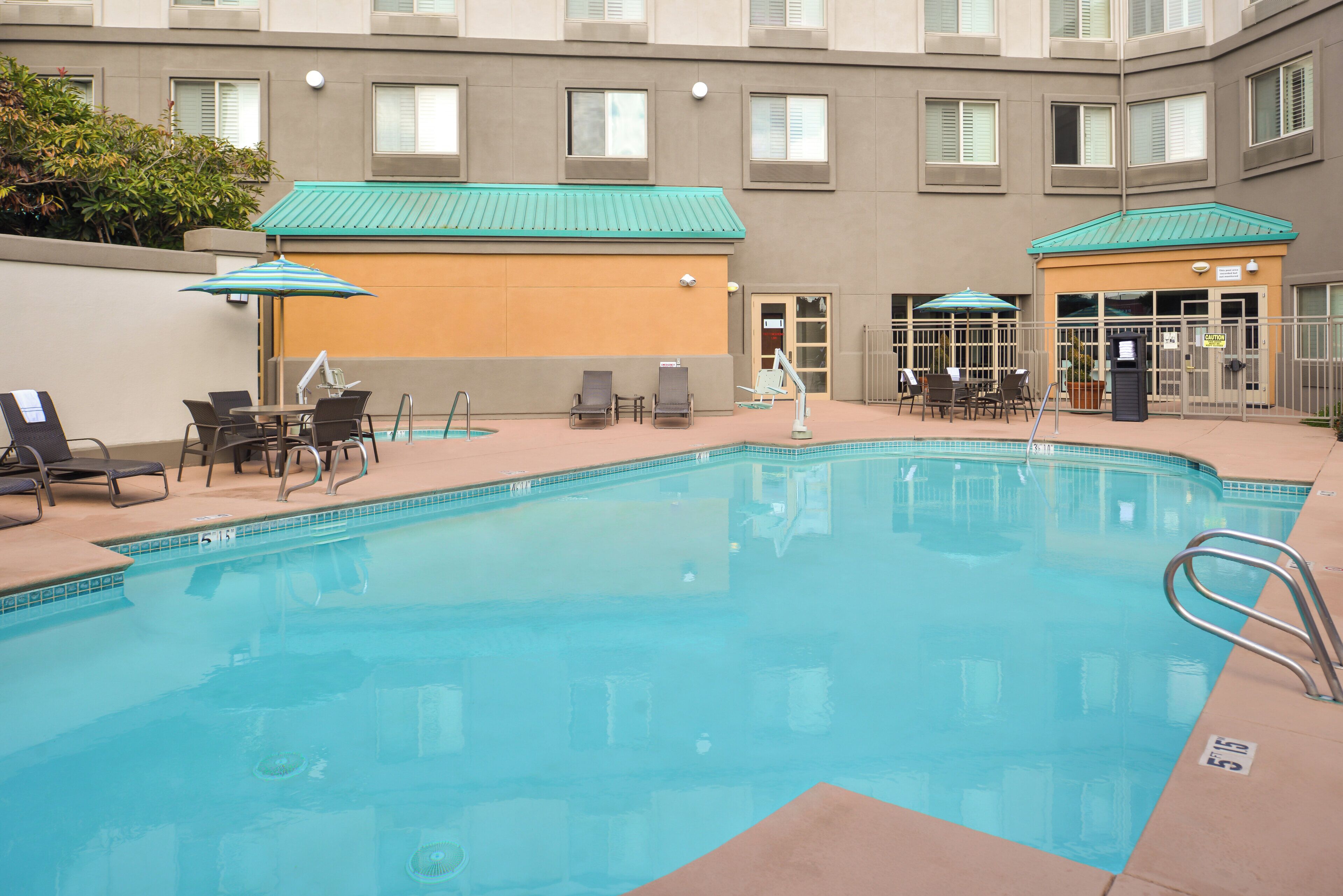 A swimming pool with lounge chairs and umbrellas, surrounded by a building with multiple windows and a door.