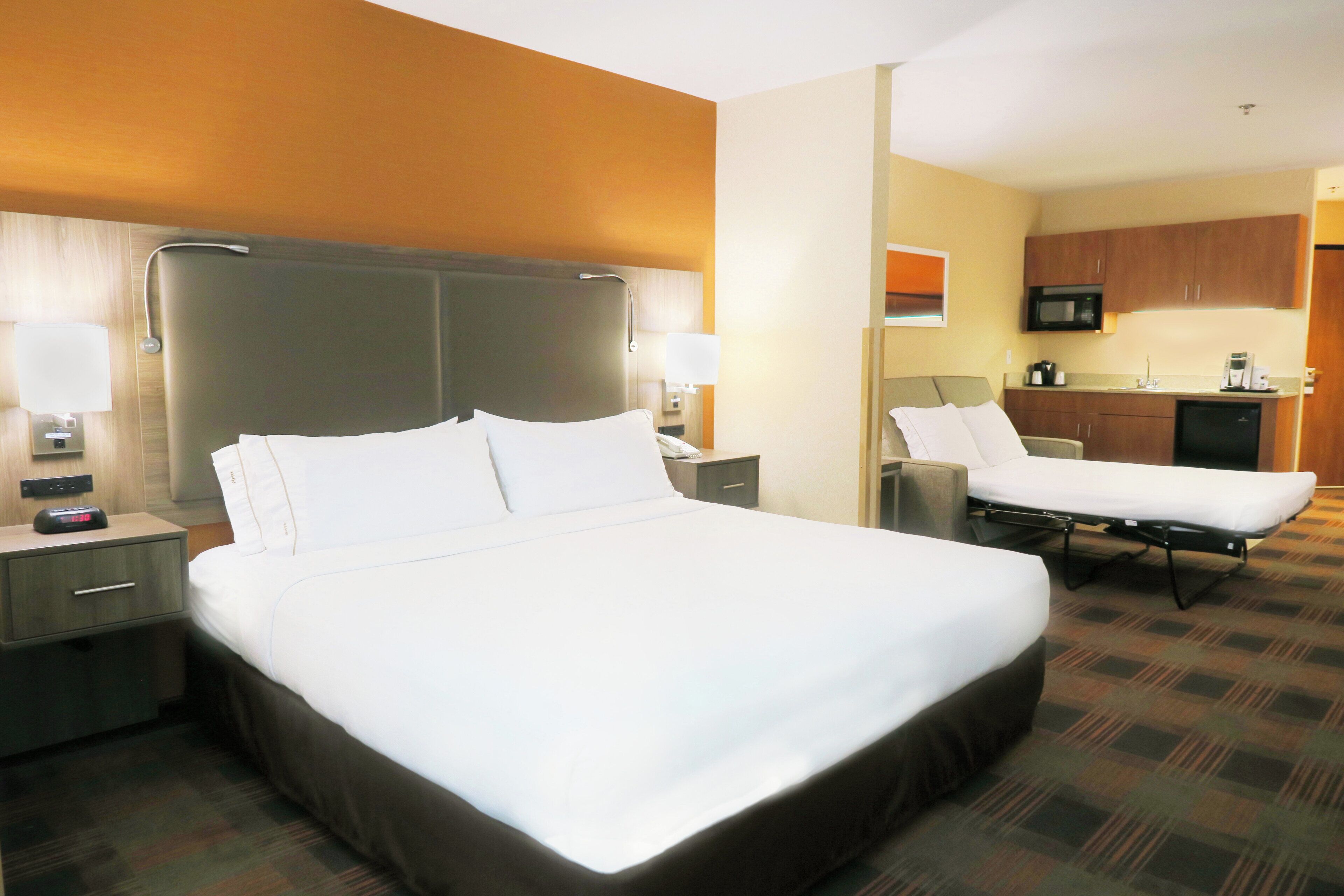 Camera d'albergo con un letto grande, un comodino, una lampada, una sveglia e vista sull'area cucina adiacente.