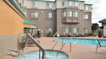 Zona de piscina de un hotel con jacuzzi, sillas de descanso y un edificio con múltiples ventanas y balcones al fondo.