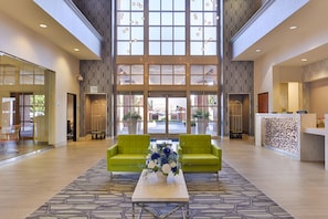 A spacious lobby with a high ceiling, large windows, a reception desk, and a green sofa with a floral arrangement.