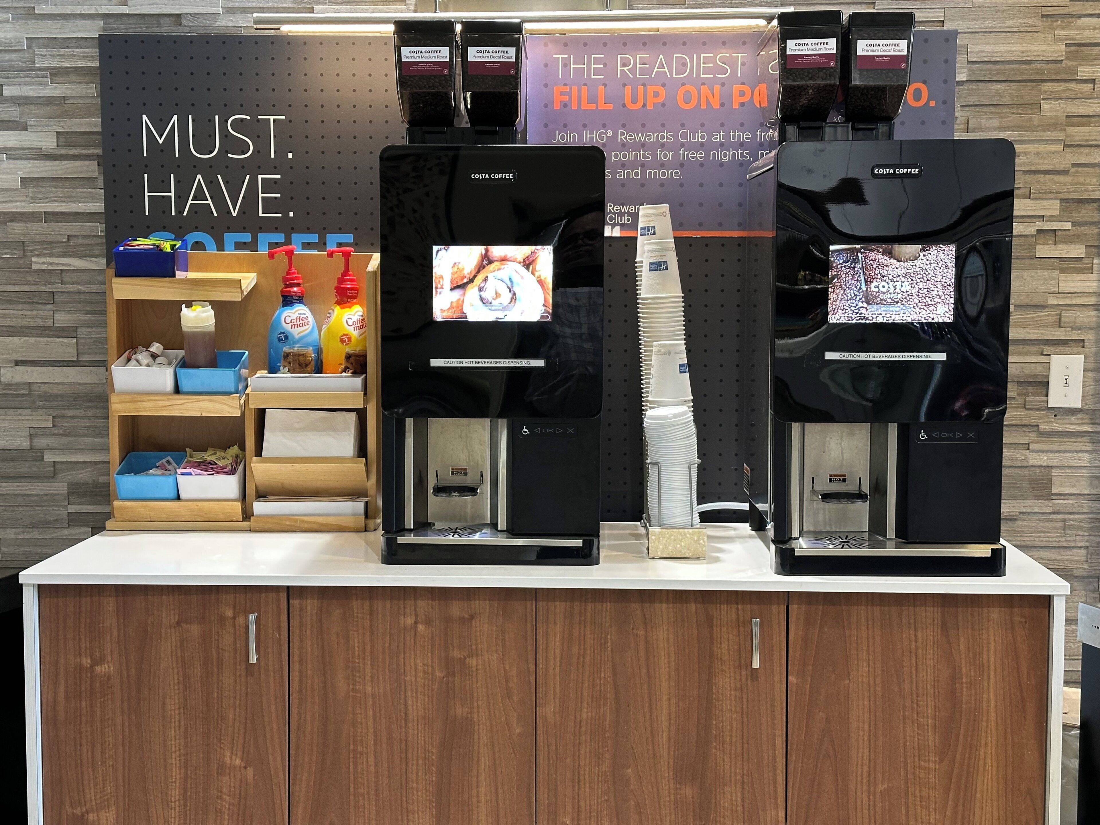 A coffee station with three coffee dispensers, a sign promoting a rewards club, and a shelf with coffee products.
