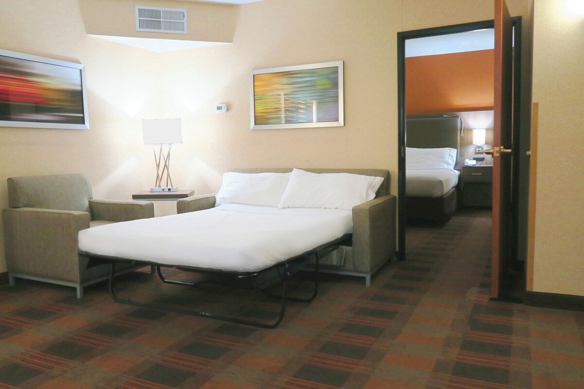 A hotel room with a bed, a chair, a bedside table, and two paintings on the wall.
