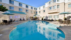 A hotel pool area with multiple lounge chairs and umbrellas, surrounded by a multi-story building with balconies.
