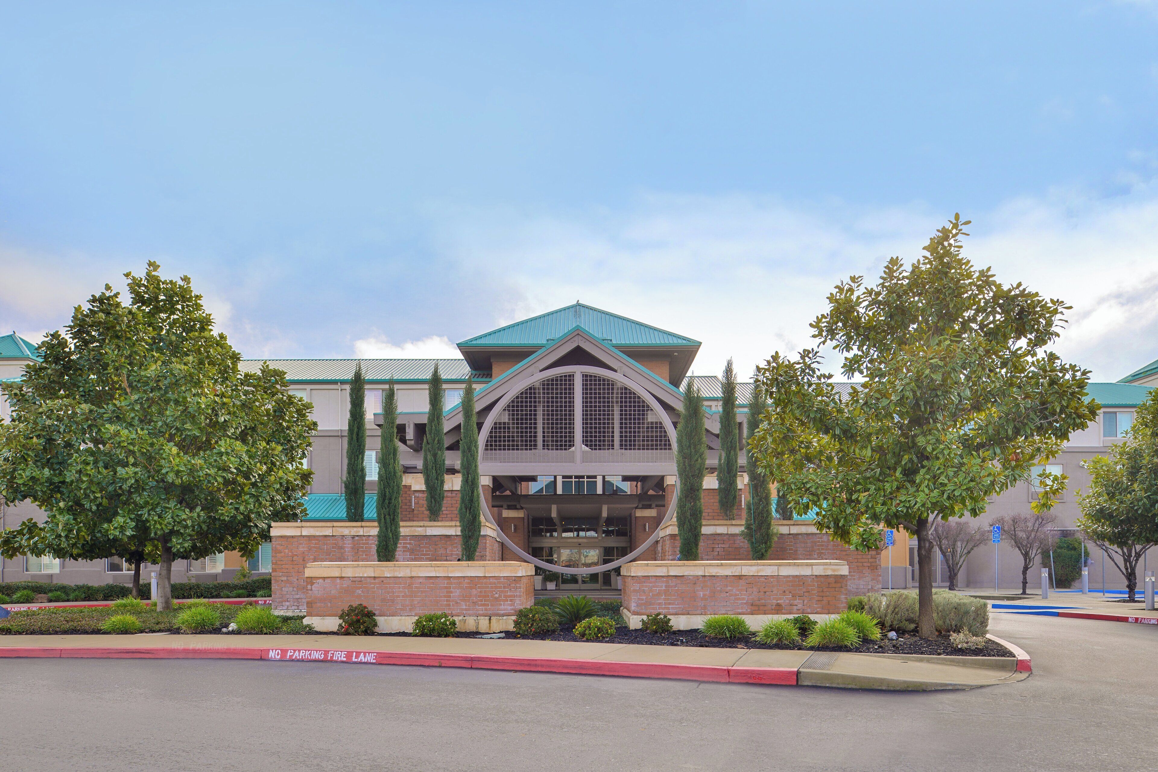 A building with a central entrance, surrounded by trees and a red curb indicating a no parking fire lane.