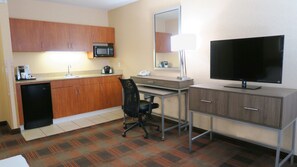 A compact hotel room with a kitchenette, a desk with a chair, a flat-screen TV, and a large mirror.