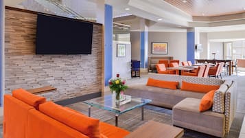 A modern lobby with a flat-screen TV, a seating area with orange sofas, a glass coffee table, and a dining area with tables and chairs.