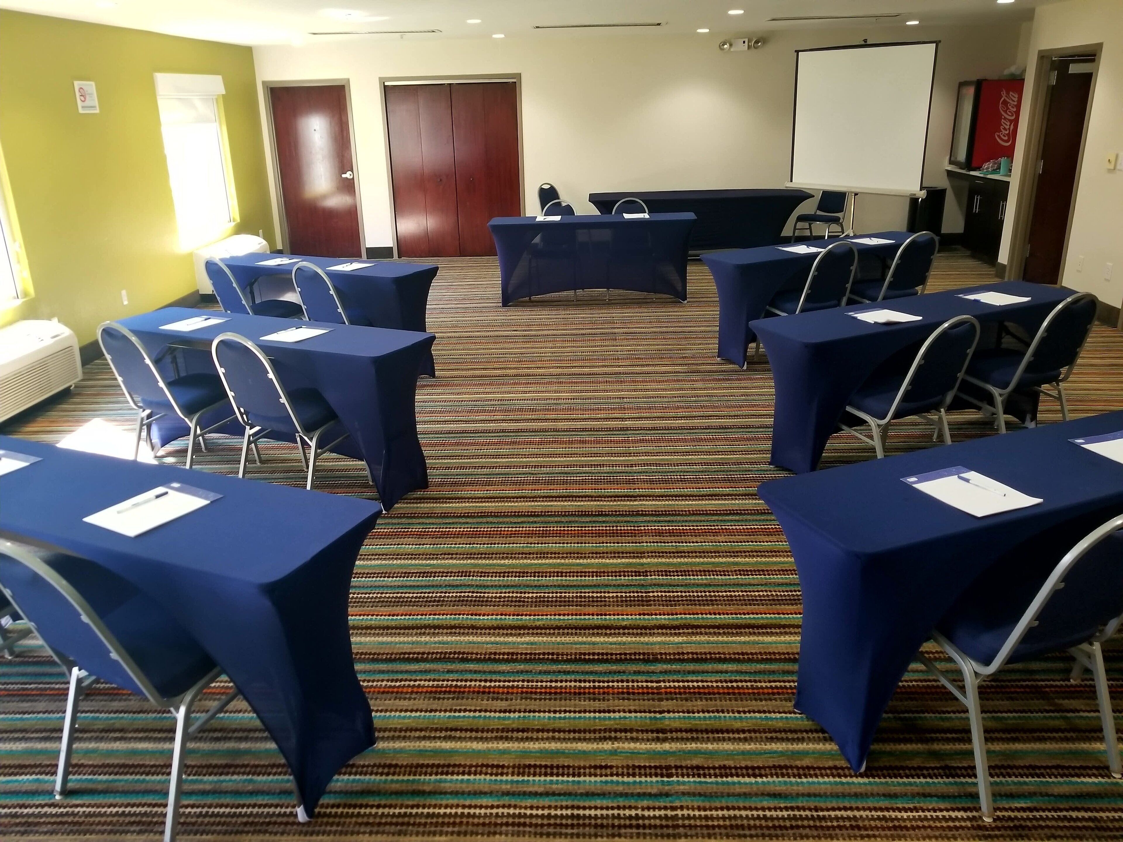 A conference room with tables and chairs, a projector screen, and a Coca-Cola refrigerator.