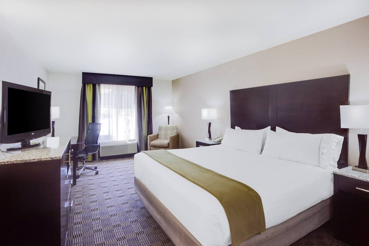 A hotel room with a large bed, a desk with a TV, a chair, and a window with curtains.