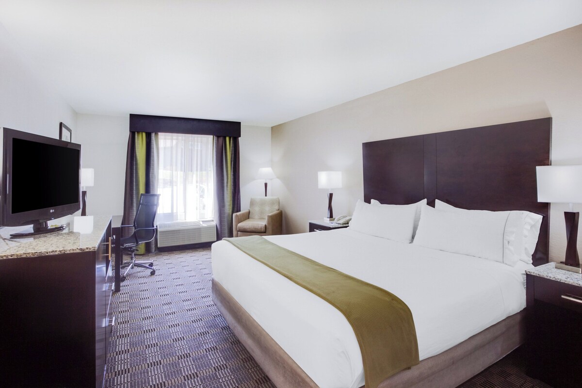 Ein Hotelzimmer mit einem großen Bett, einem Schreibtisch mit Fernseher, einem Sessel und einem Fenster mit Vorhängen.