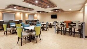 A spacious restaurant with multiple tables and chairs, a flat-screen TV mounted on the wall, and wooden ceiling panels.