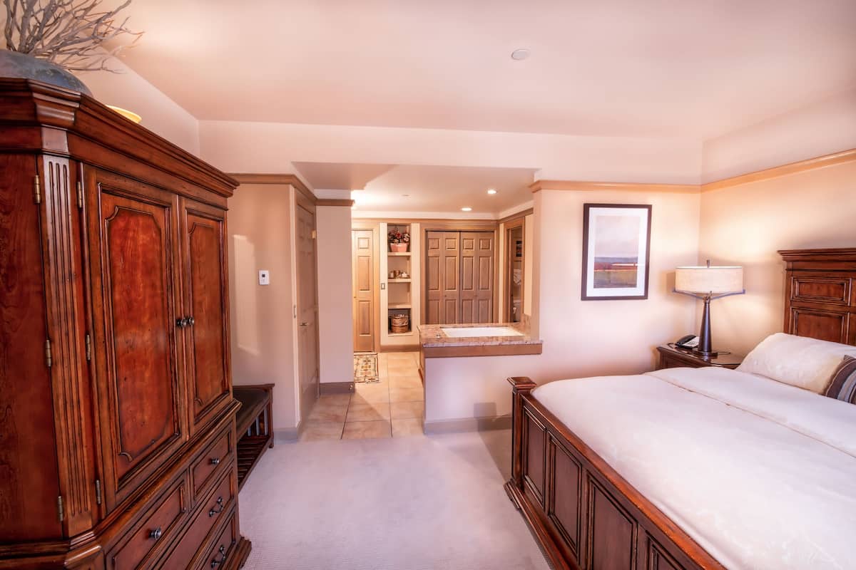 Un dormitorio con un amplio armario de madera, una cama con cubrecama blanco, una mesita de noche con lámpara y un baño con lavabo y espejo.