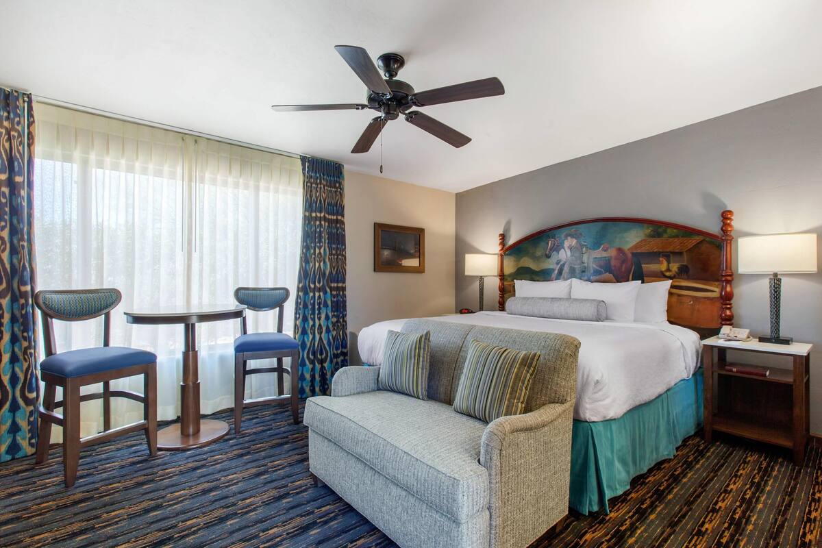 A hotel room with a bed, a sofa, two chairs, a ceiling fan, a painting, a nightstand, and a lamp.