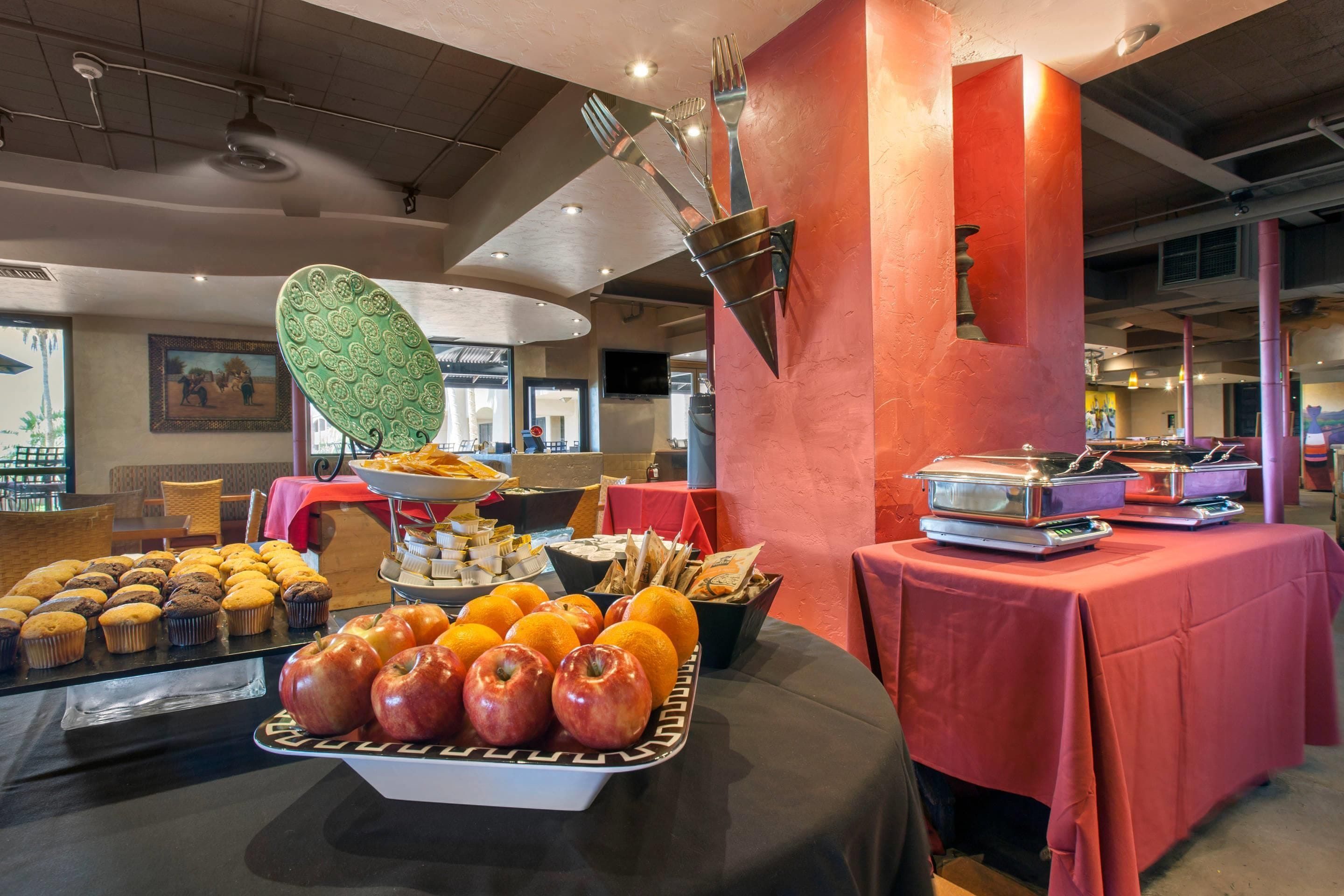 A buffet setup with various food items, including apples and muffins, and a decorative wall with a unique design.