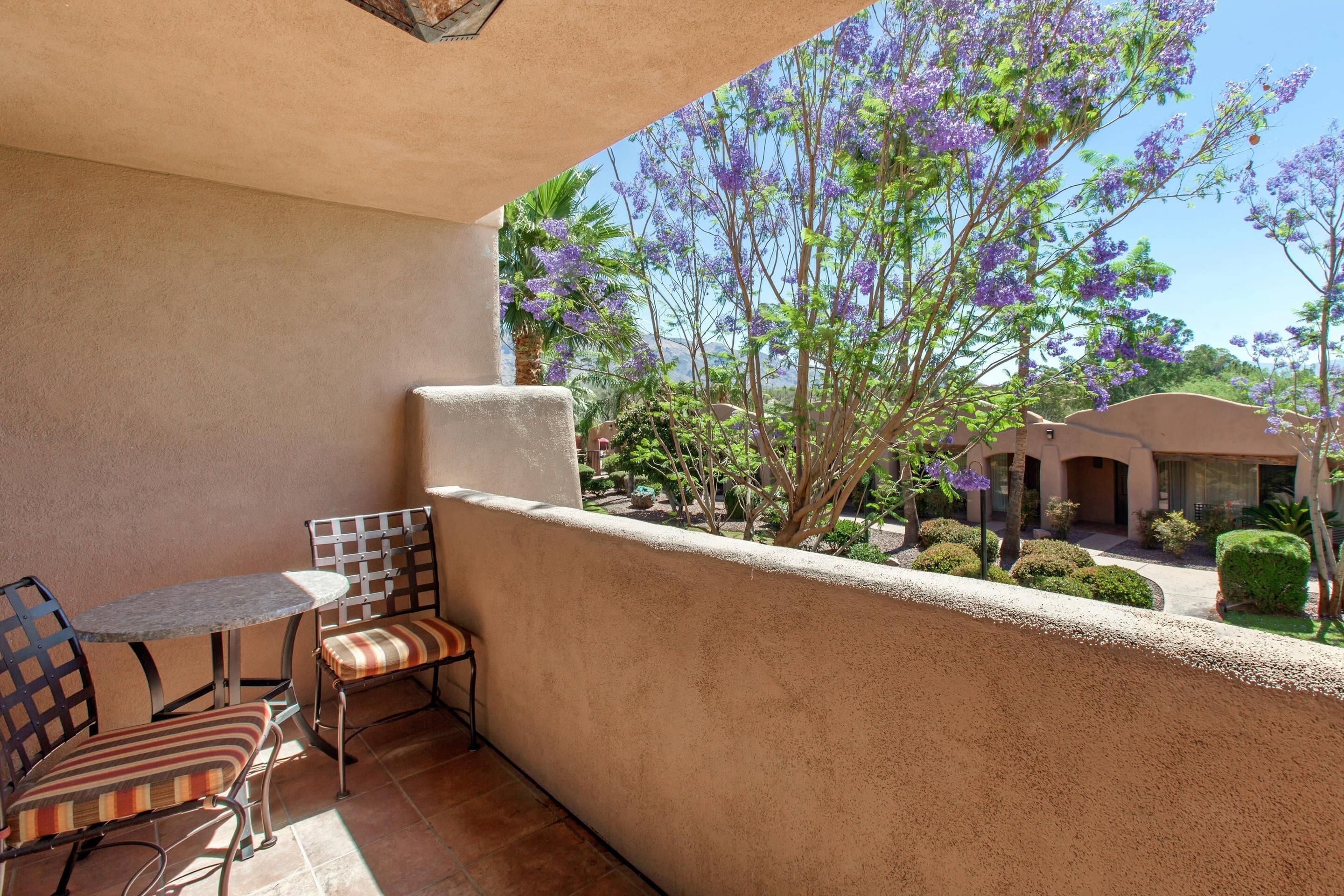 A balcony with a table and chairs, a view of purple flowering trees, and a building in the background.
