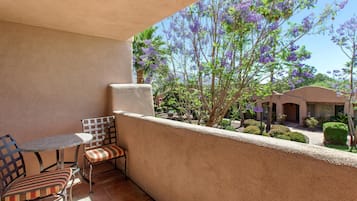 A balcony with a table and chairs, a view of purple flowering trees, and a building in the background.