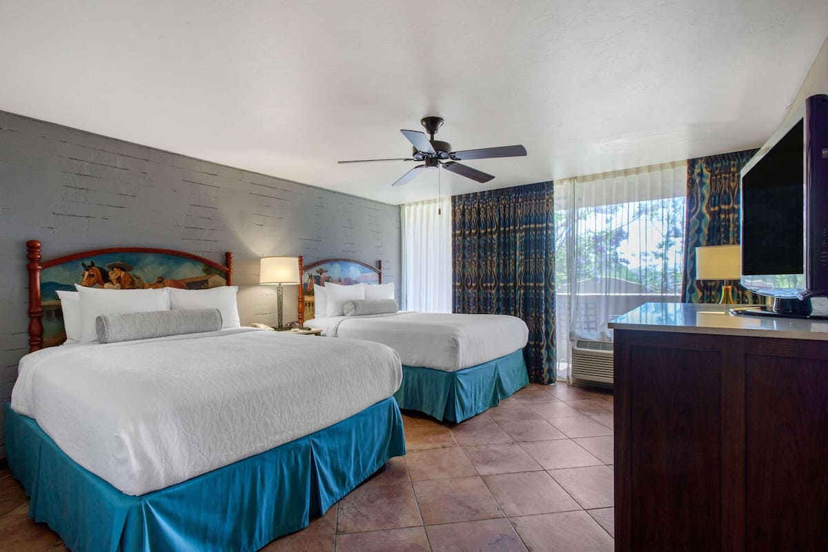 A hotel room with two beds, a ceiling fan, a desk with a computer, and a large window with curtains.