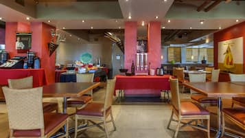 Restaurante com várias mesas e cadeiras, área de buffet e balcão com alimentos.