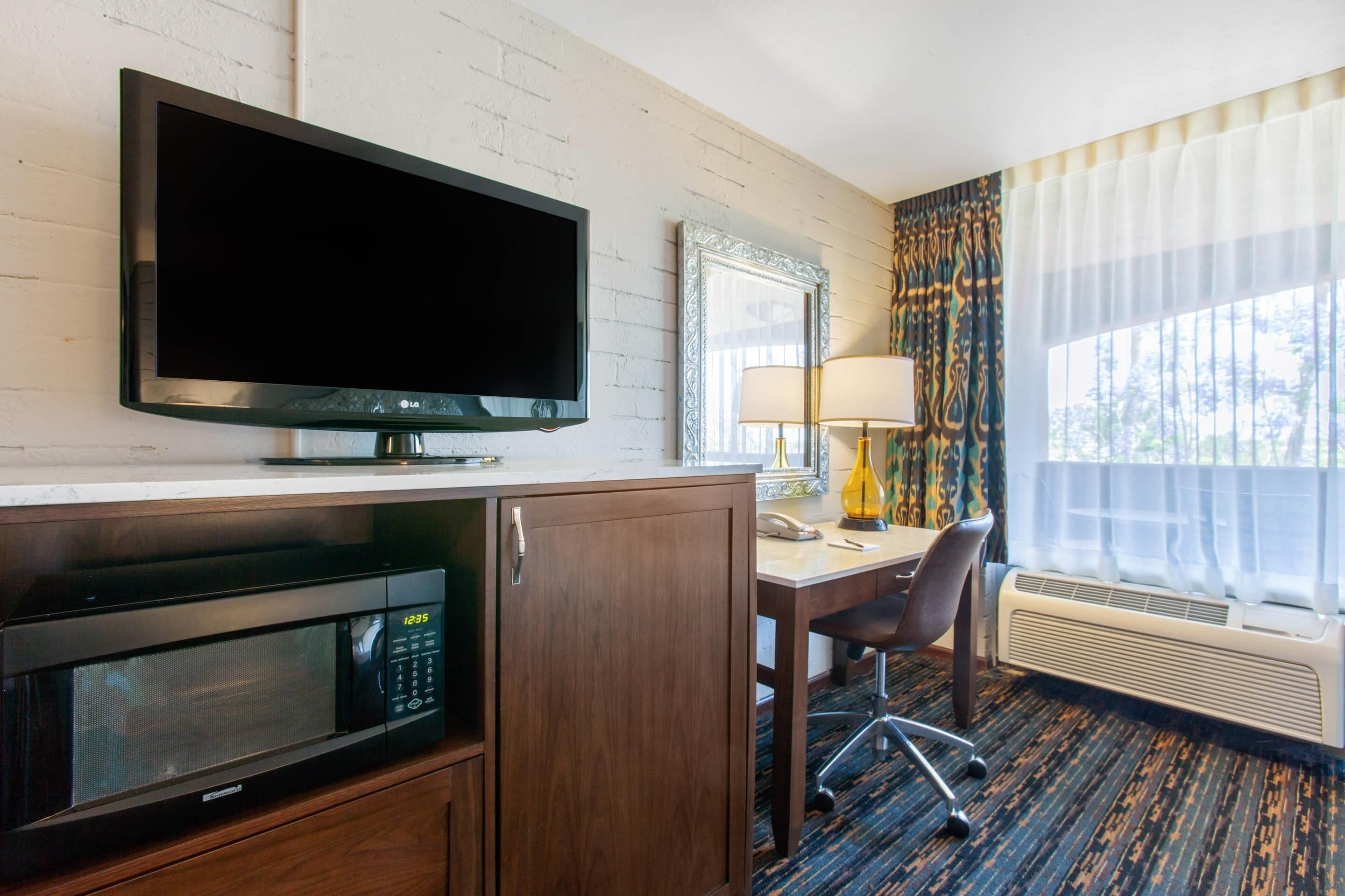 Une chambre d’hôtel équipée d’une télévision à écran plat, d’un four à micro-ondes, d’un bureau avec une chaise, d’une lampe et d’une grande fenêtre avec des rideaux.