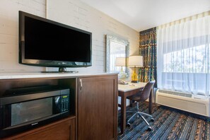 A hotel room with a flat-screen TV, a microwave, a desk with a chair, a lamp, and a large window with curtains.