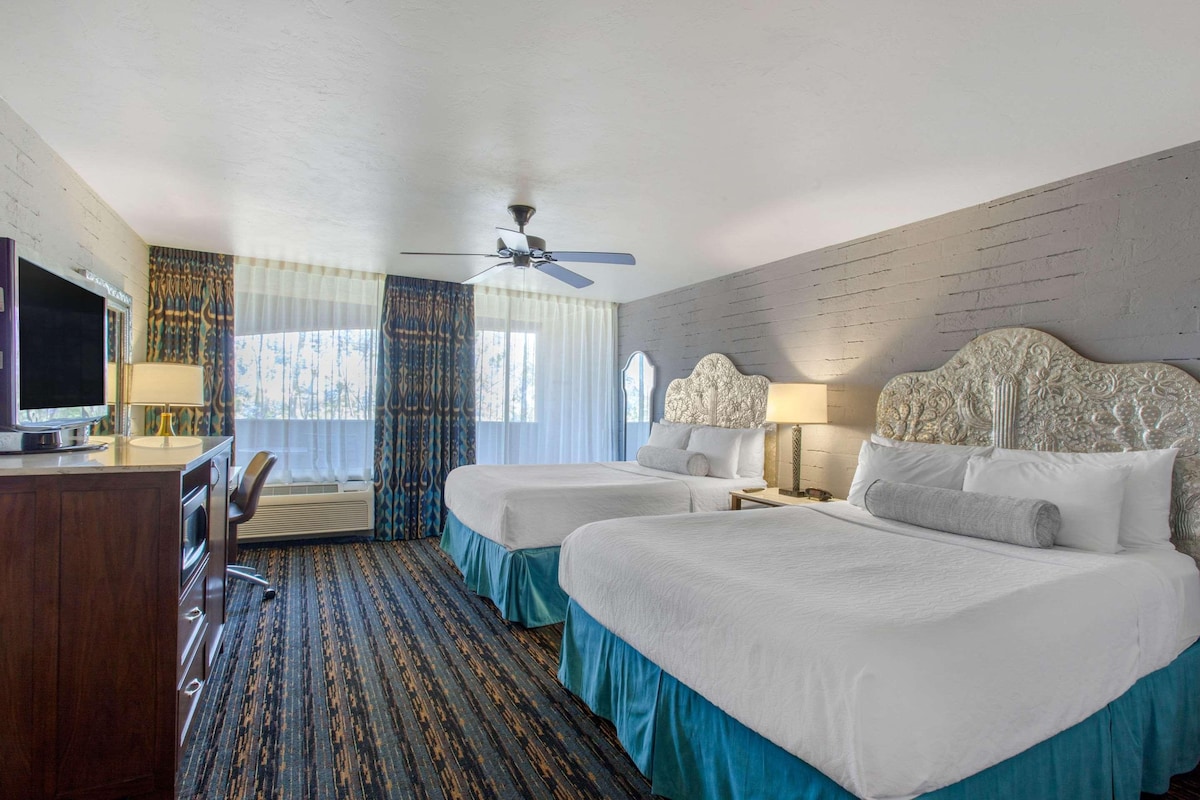 A hotel room with two beds, a desk with a TV, a ceiling fan, and a window with curtains.