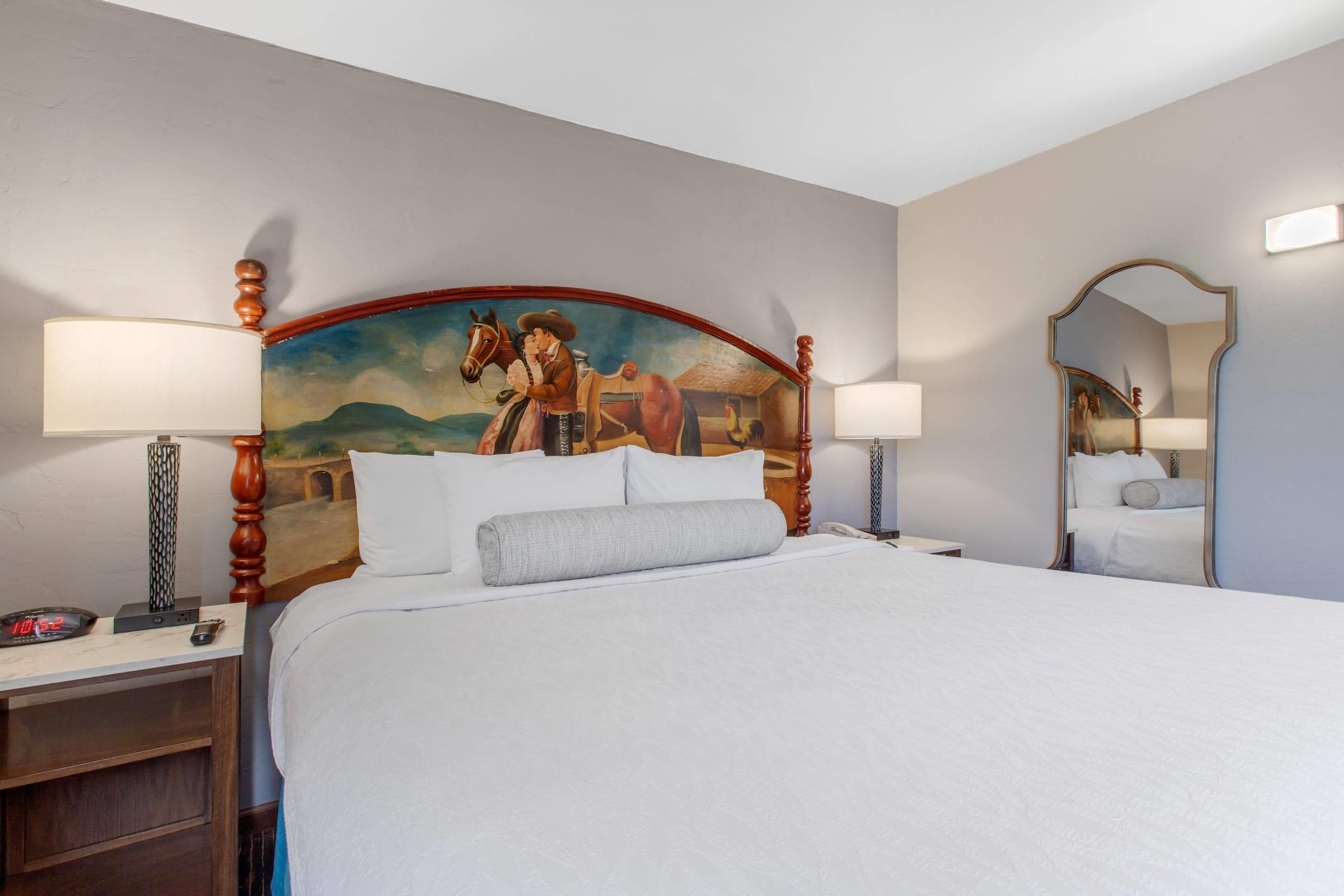 A hotel room with a large bed, two bedside lamps, a nightstand, a digital clock, a mirror, and a wall-mounted painting.
