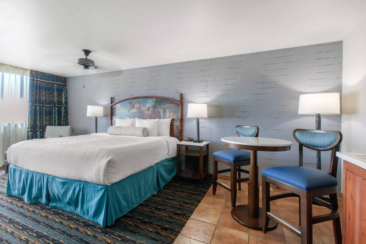 A hotel room with a bed, two chairs, a table, a lamp, and a ceiling fan.