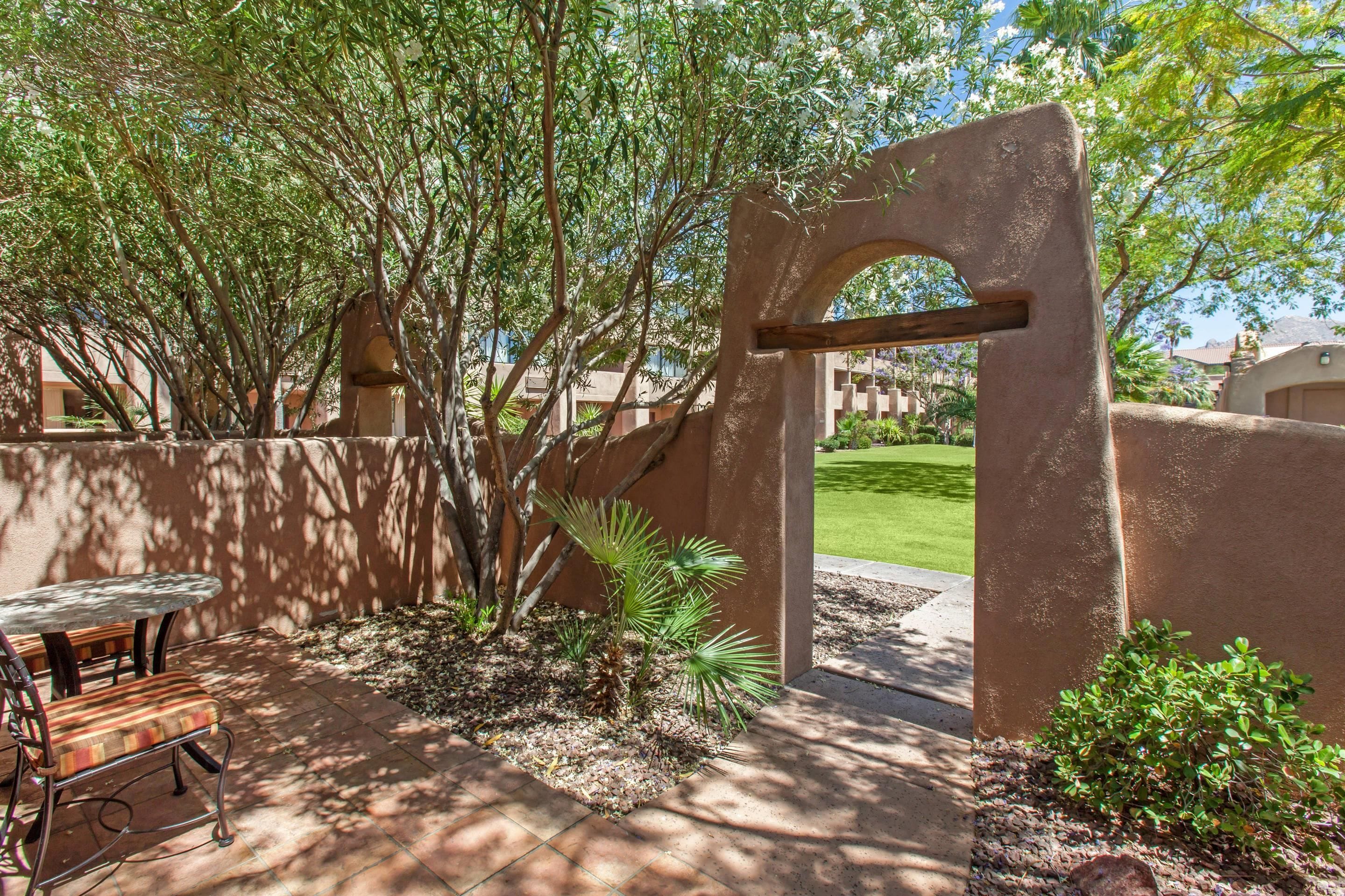 An outdoor patio area with a table and chairs, surrounded by a low wall and various plants.