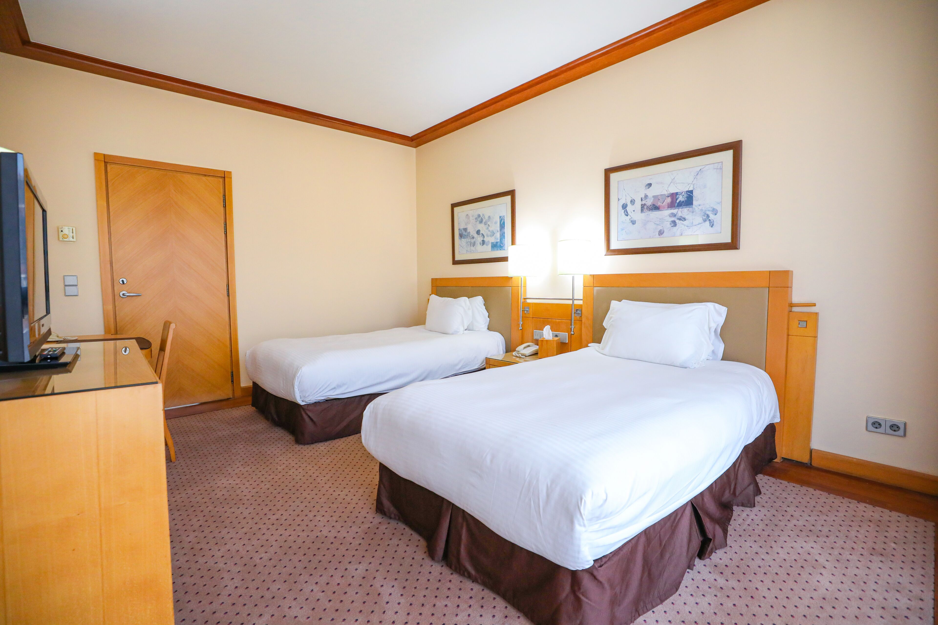 A hotel room with two single beds, a desk with a computer, a door, and two framed pictures on the wall.