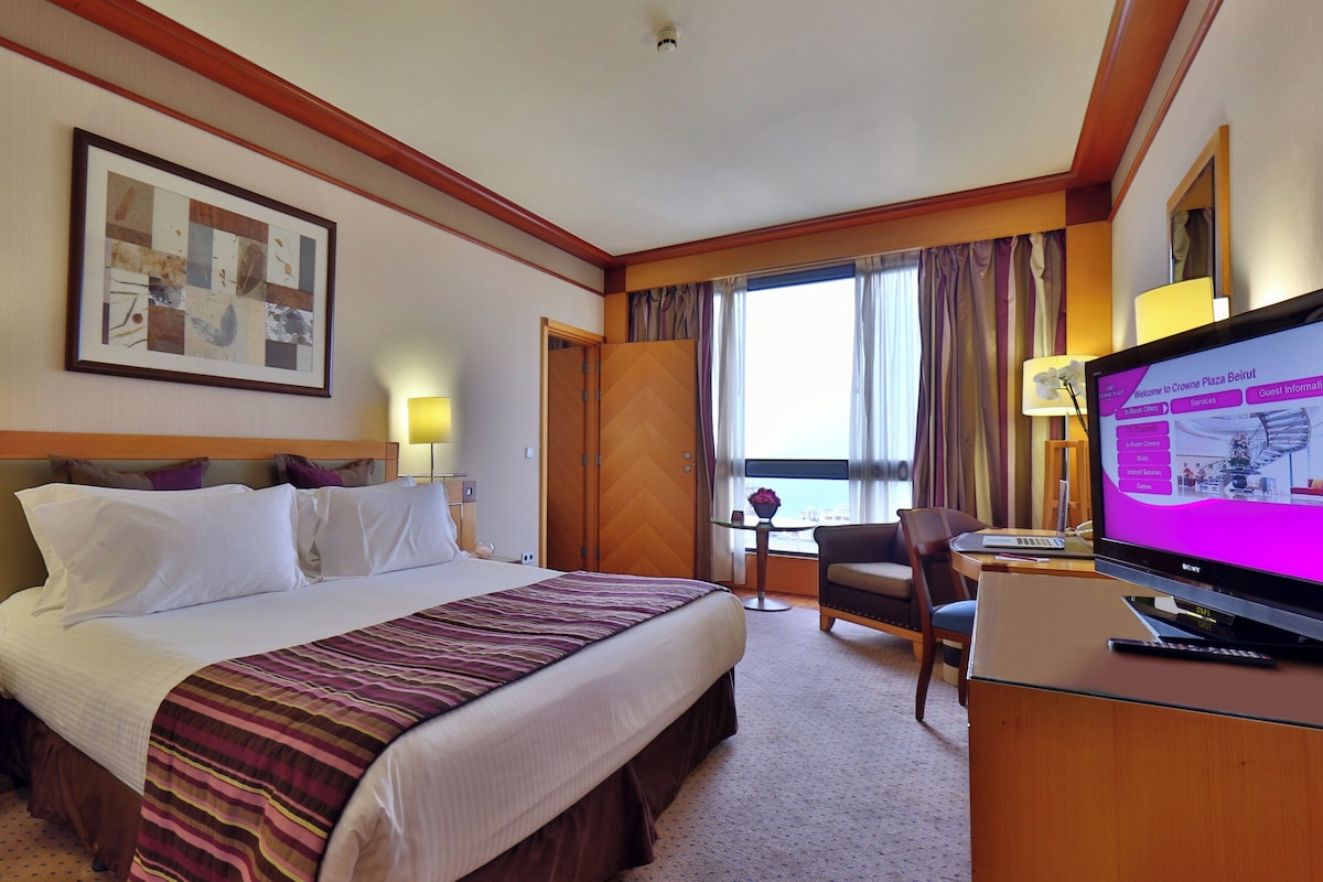 A hotel room with a bed, bedside tables, a television, a desk, and a window with curtains.