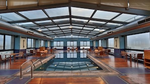 A spacious indoor swimming pool with a glass ceiling, surrounded by wooden tables and chairs, and large windows offering views of the city. - Crowne Plaza Hotel Beirut by IHG (Beirut)
