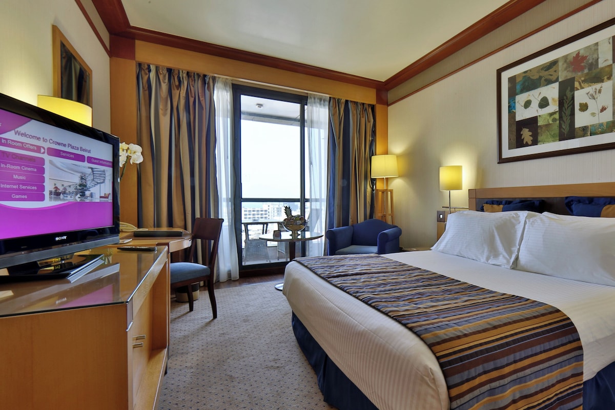 A hotel room with a bed, a desk with a TV, a chair, a balcony view, and a painting on the wall.