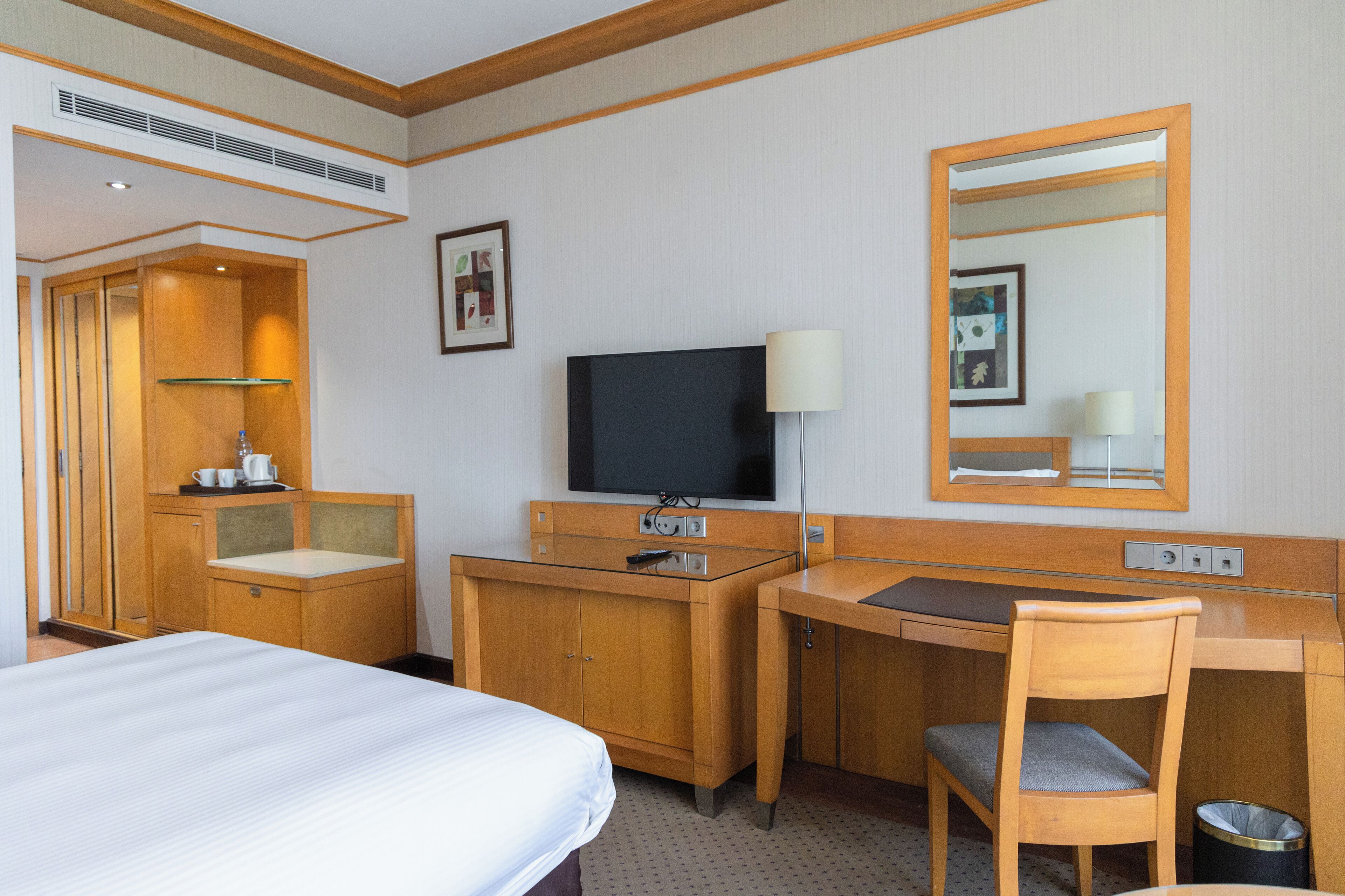 A hotel room with a bed, a desk with a chair, a television, a mirror, a small cabinet, and a trash bin.