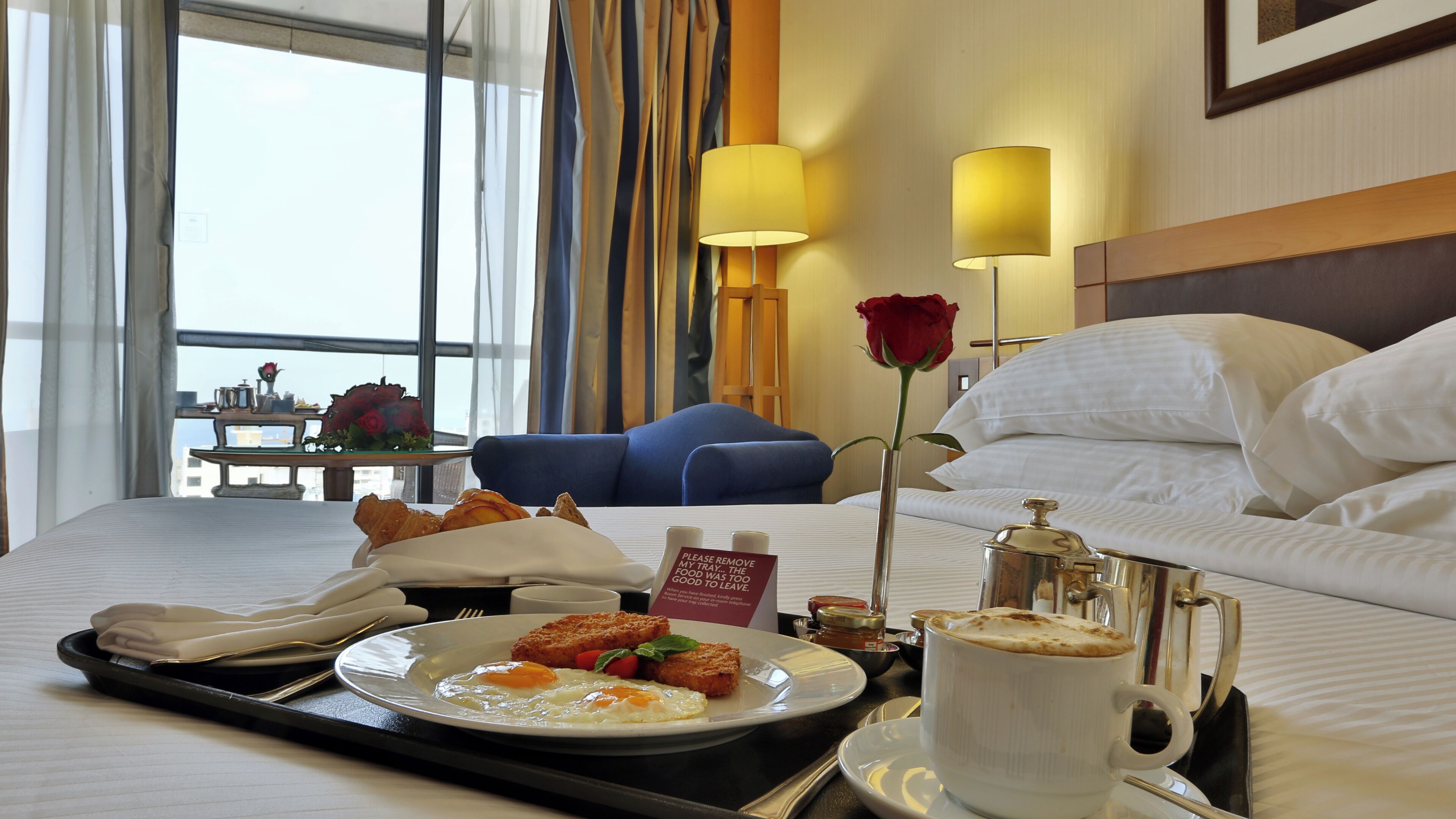 A hotel room with a bed, a tray with breakfast, a coffee cup, a rose, and a view of the city.