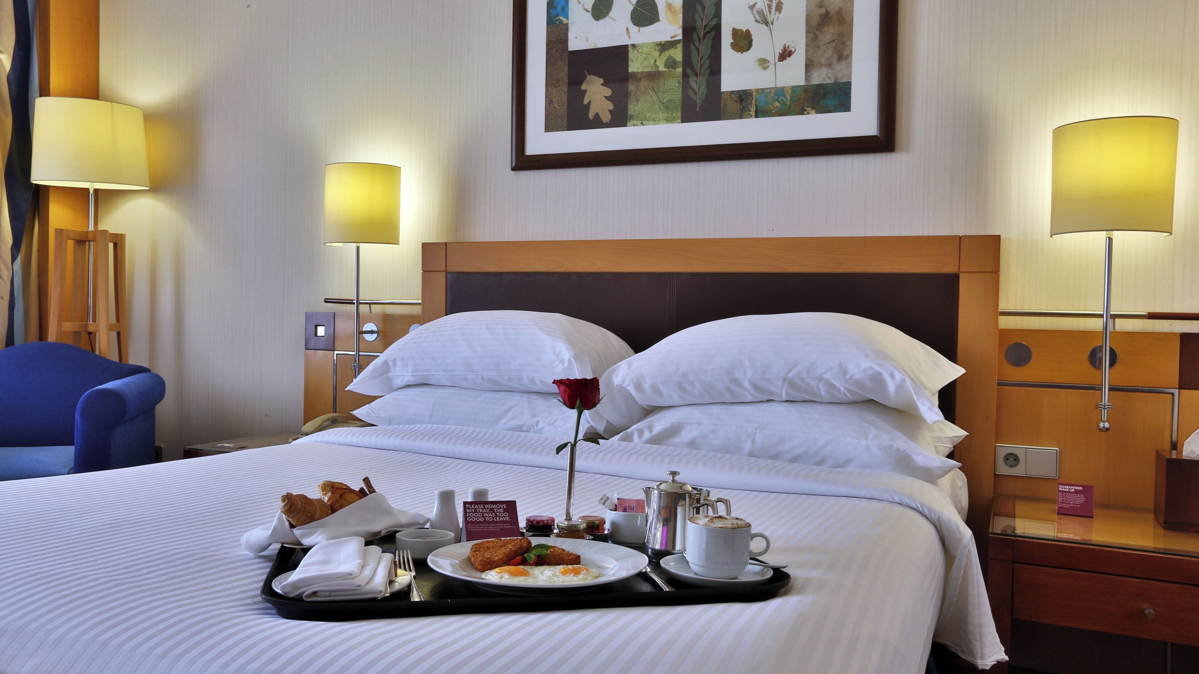 A hotel room with a neatly made bed, a tray with breakfast items, a single rose on the pillow, and a framed botanical artwork on the wall.