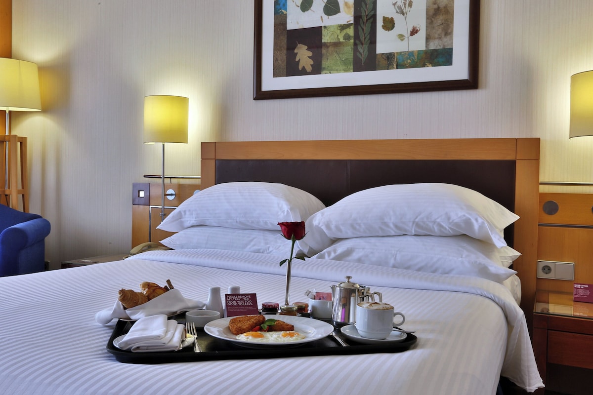 A hotel room with a neatly made bed, a tray with breakfast items, a single rose on the pillow, and a framed botanical artwork on the wall.