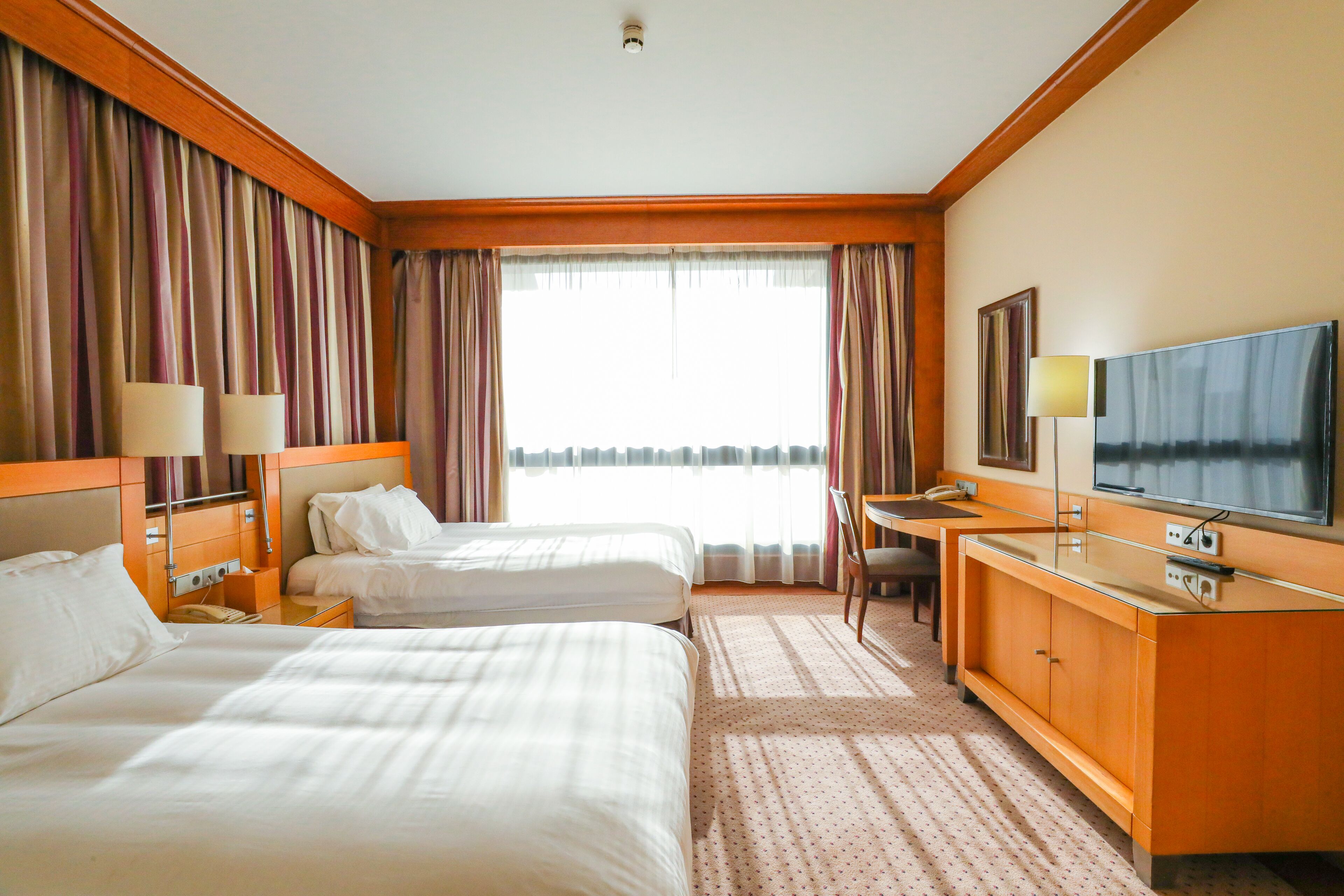A hotel room with two beds, a desk, a television, and a window with curtains.