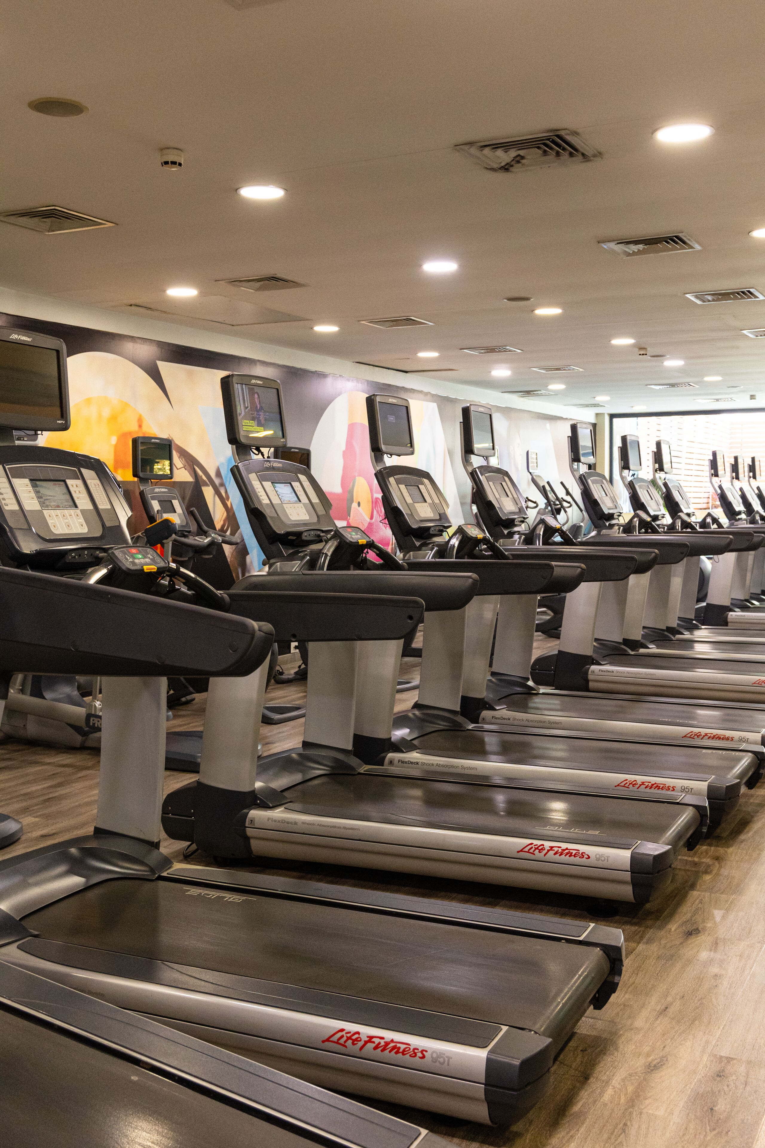 Une rangée de tapis de course dans une salle de sport, avec un plancher en bois et une fresque murale.