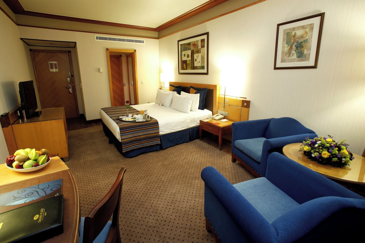 A hotel room with a bed, a desk with a computer, a chair, a sofa, and a dining table with fruit.