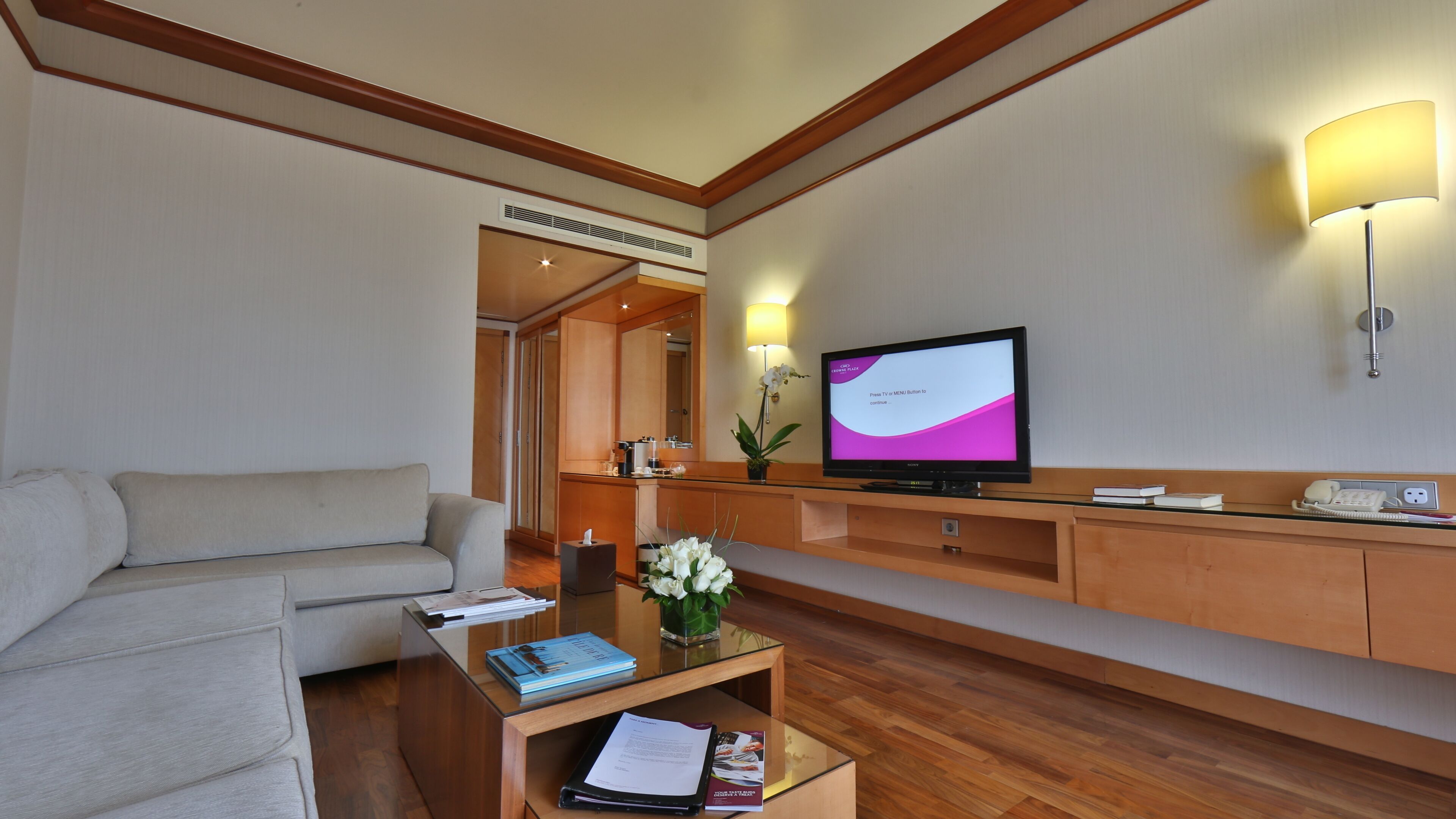 A hotel room with a sofa, a coffee table with books and a vase of flowers, a TV on a stand, a bedside table with a lamp, and a wooden cabinet.