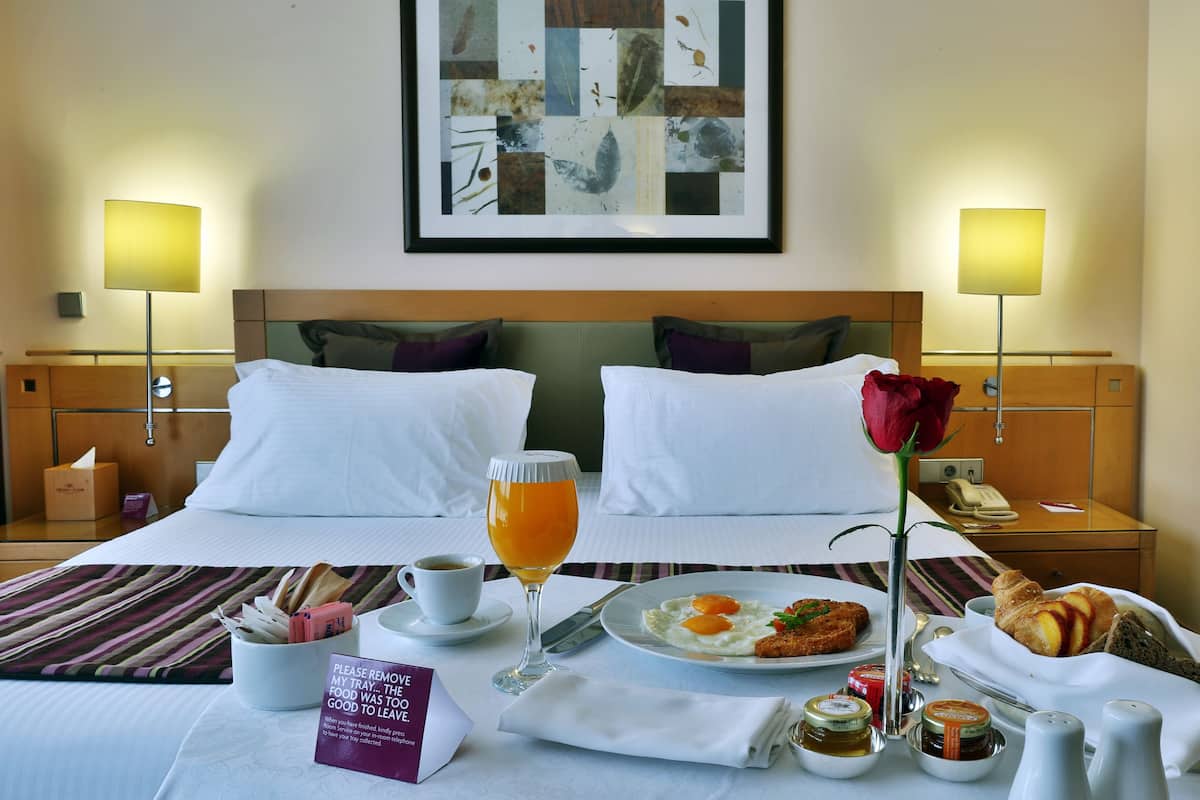 Quarto de hotel com cama bem arrumada, bandeja de café da manhã com ovos, torradas e um croissant, um vaso com uma única rosa vermelha e uma obra de arte abstrata emoldurada na parede.