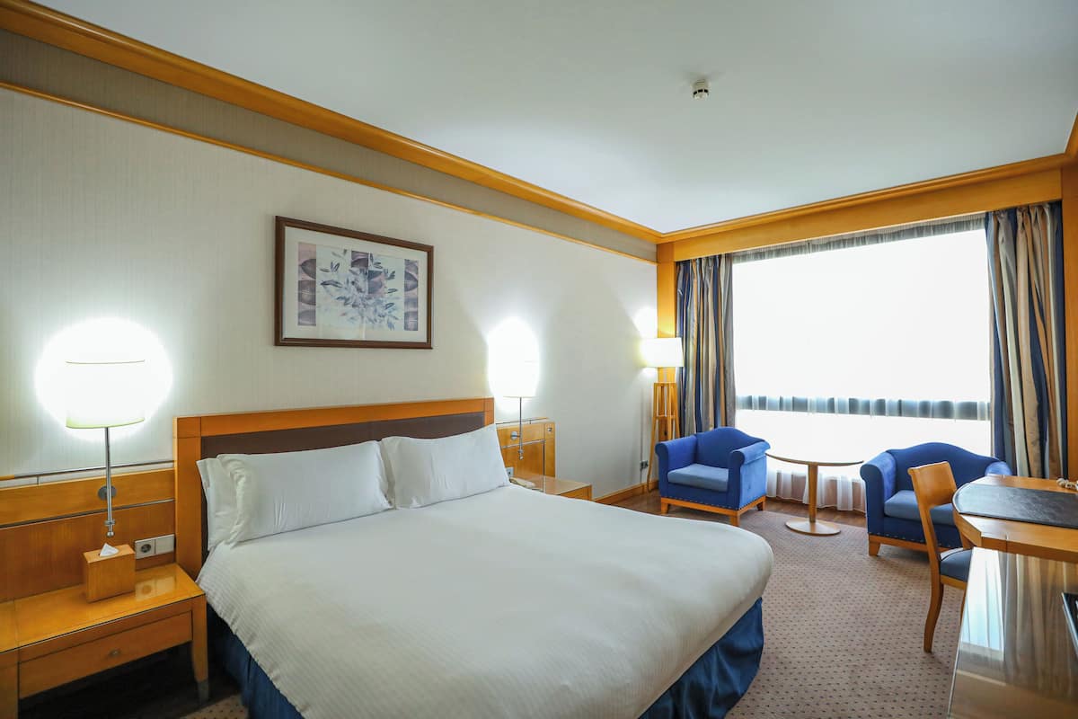 Quarto de hotel com uma cama grande, duas mesas de cabeceira, uma escrivaninha com cadeira, uma pequena mesa com duas cadeiras e uma janela grande com cortinas.