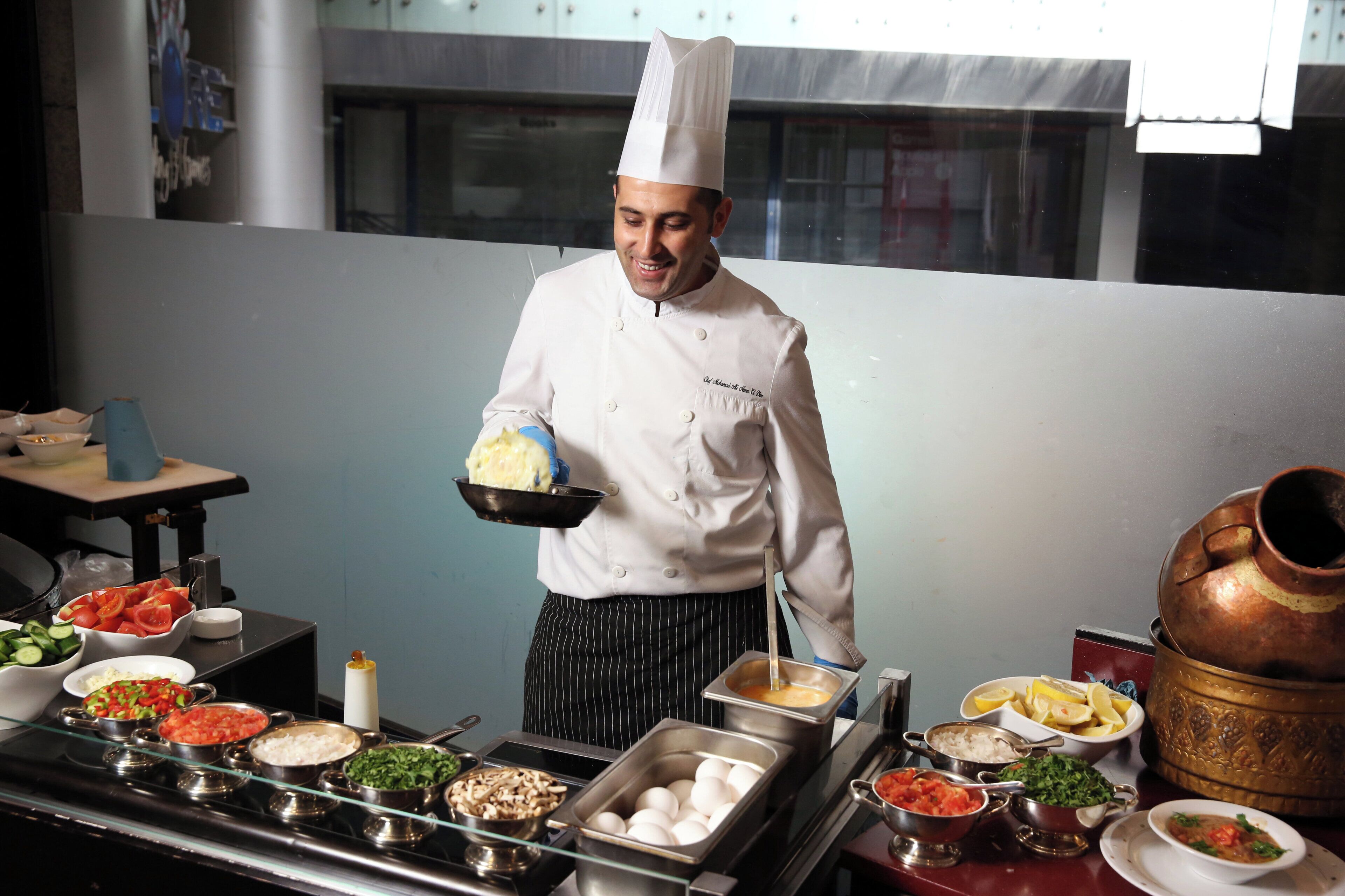 Un chef se tient dans une cuisine, tenant un bol rempli de nourriture. On peut voir divers aliments et ustensiles de cuisine sur le plan de travail.