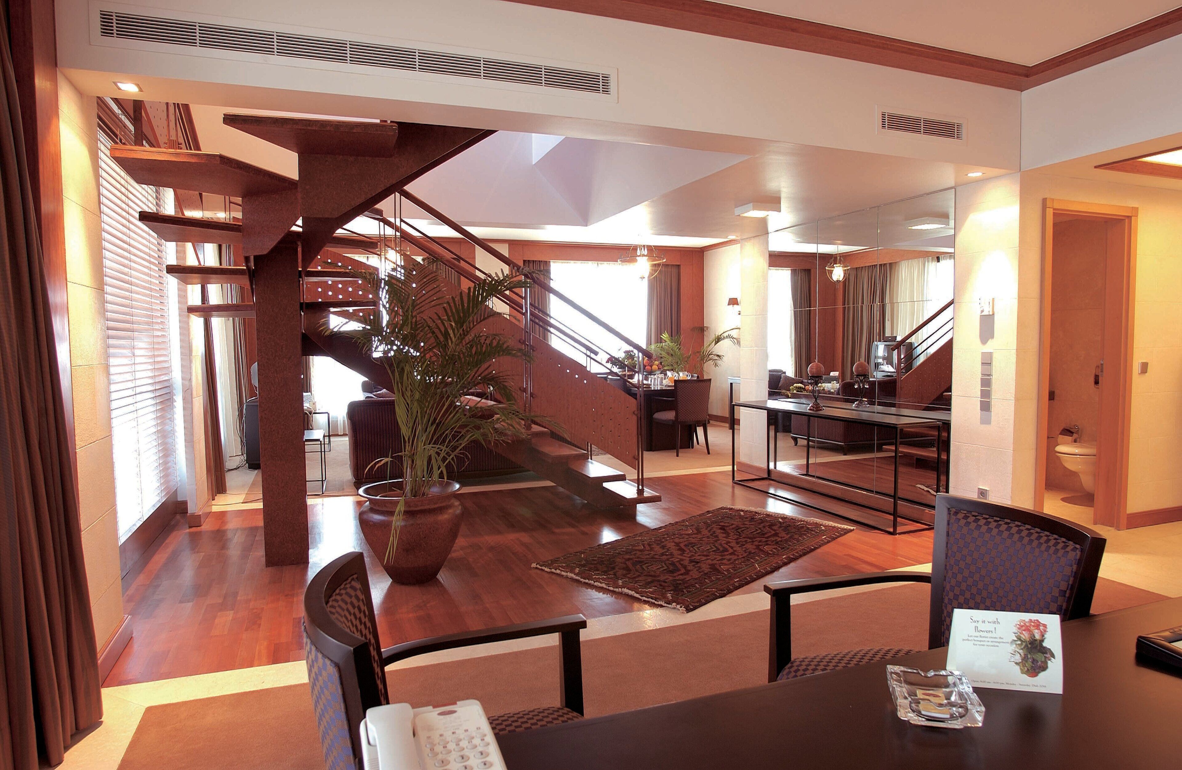 Um amplo hall de hotel com escadaria de madeira, área de refeições e balcão de recepção.