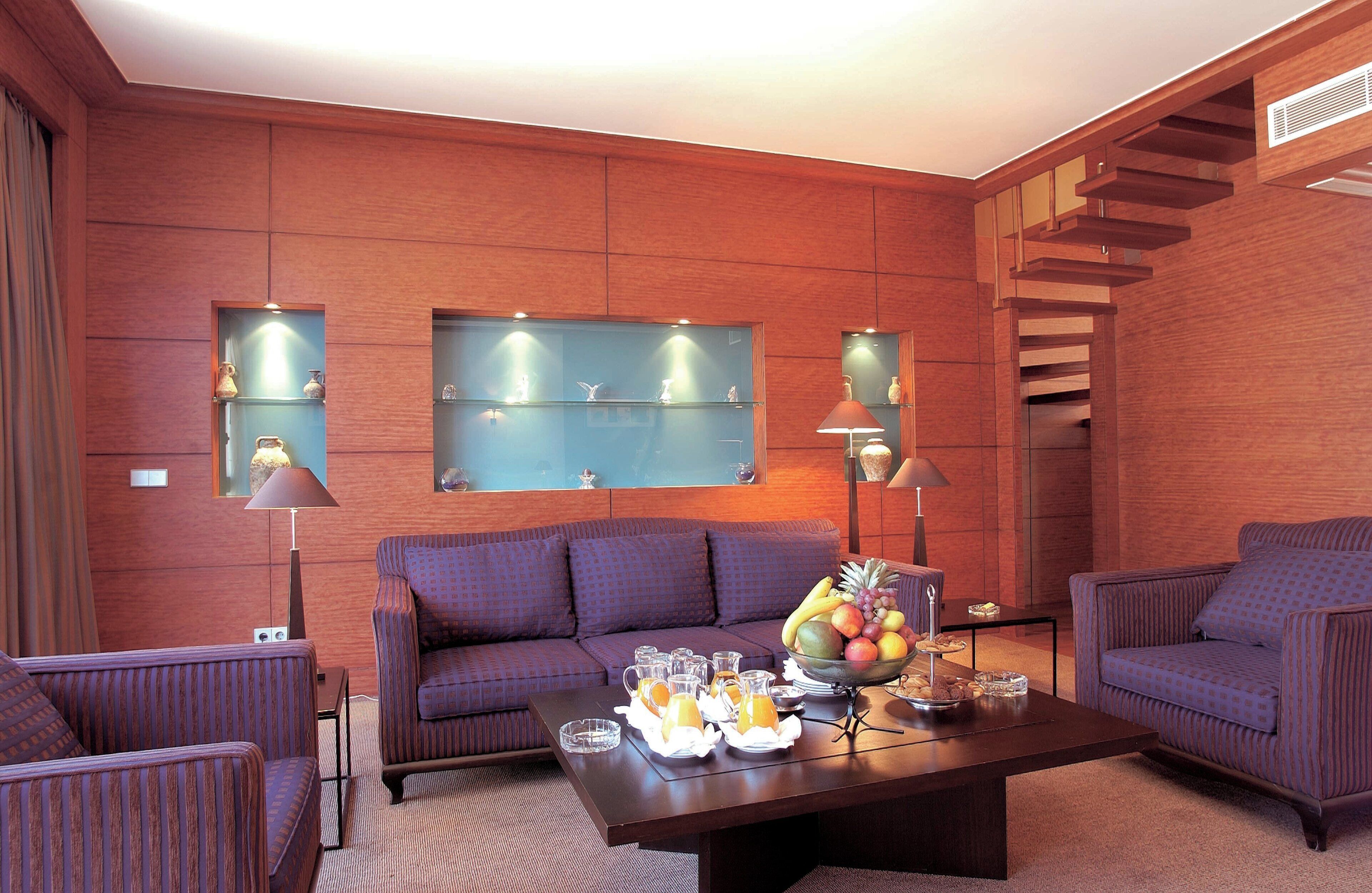 A modern living room with a sofa, armchairs, a coffee table with fruit and drinks, and wall-mounted shelves with decorative items.